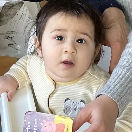 En familj sitter tillsammans i ett vardagsrum. En vuxen och ett barn leker vid en grön leksak. I bakgrunden syns en beige soffa och ett ljust trägolv.