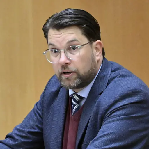 Tre personer står vid talarstolar i en politisk debatt. De är klädda i formella kläder, inklusive en blå kavaj, en ljus kavaj och en mörk kostym. Mikrofoner och ett glas vatten syns på talarstolarna.