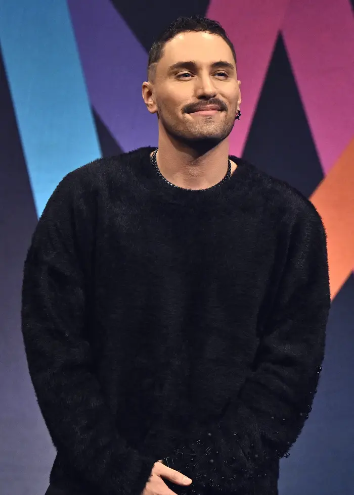 En person står på en scen framför en färgstark logotyp för Melodifestivalen 2026. Personen är klädd i svart och poserar inför pressen vid en presentation av årets tävlande artister.