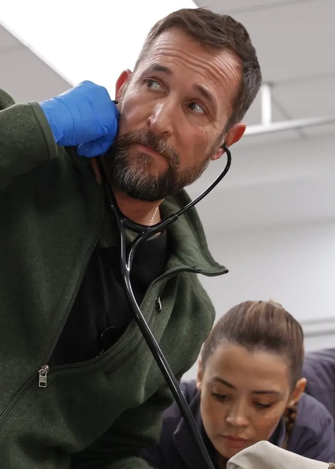 En grupp människor är samlade i ett sjukhusliknande rum med fokus på ett pågående arbete. En person i grönt och blåa handskar verkar vara involverad i en medicinsk procedur.