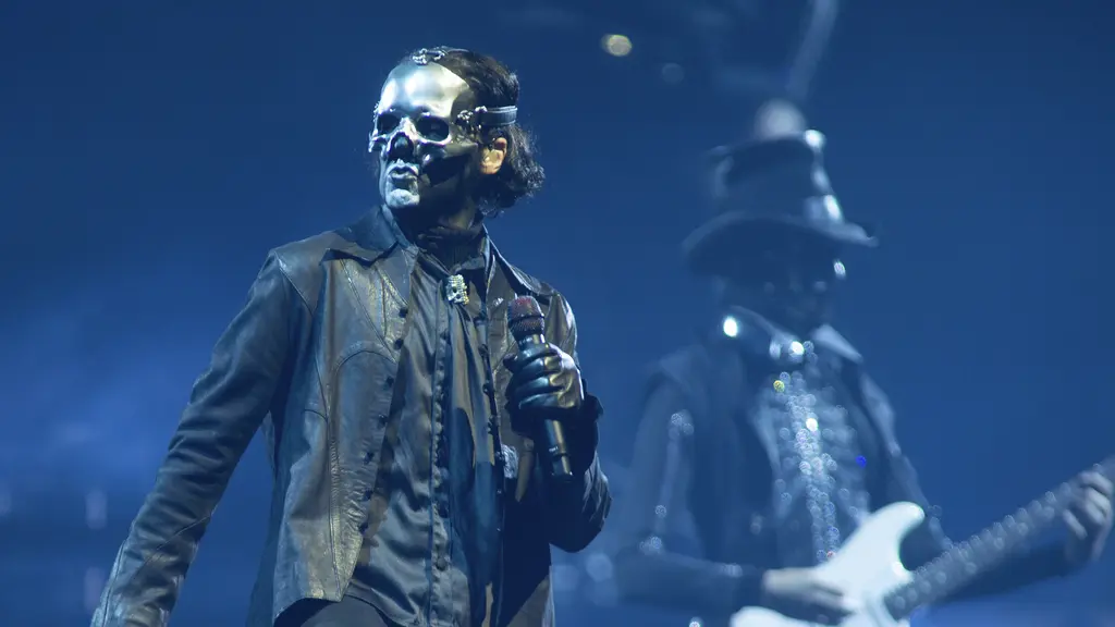 En artist i en mörk scenkostym uppträder på scen med en mikrofon i handen. Bakom honom syns en gitarrist i en liknande outfit. Scenen är upplyst med blått och violett ljus, vilket skapar en dramatisk stämning.