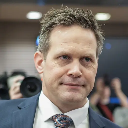 En man med glasögon och kostym samt kortklippt hår sitter i en rättssal och samtalar med en annan person i kostym. Grå väggar syns i bakgrunden.