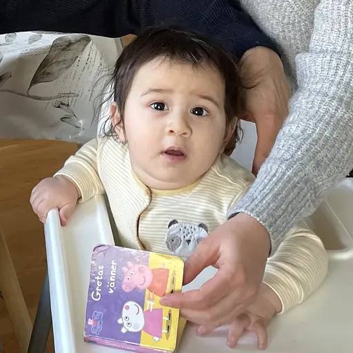 En familj sitter tillsammans i ett vardagsrum. En vuxen och ett barn leker vid en grön leksak. I bakgrunden syns en beige soffa och ett ljust trägolv.