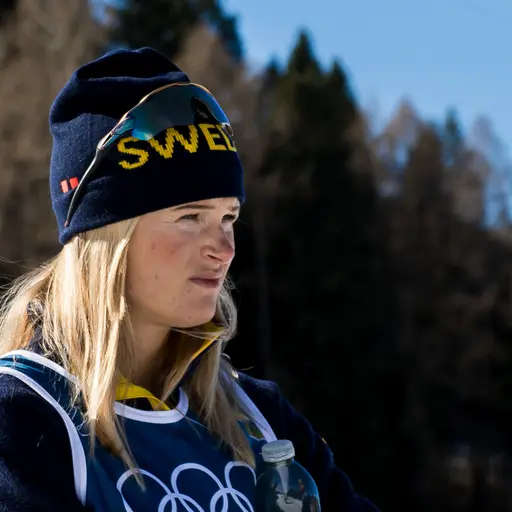 En längdskidåkare i gul och blå tävlingsdräkt håller stavar och har på sig vita handskar med Swix-logga. Personen deltar i vinter-OS i Milano Cortina 2026.