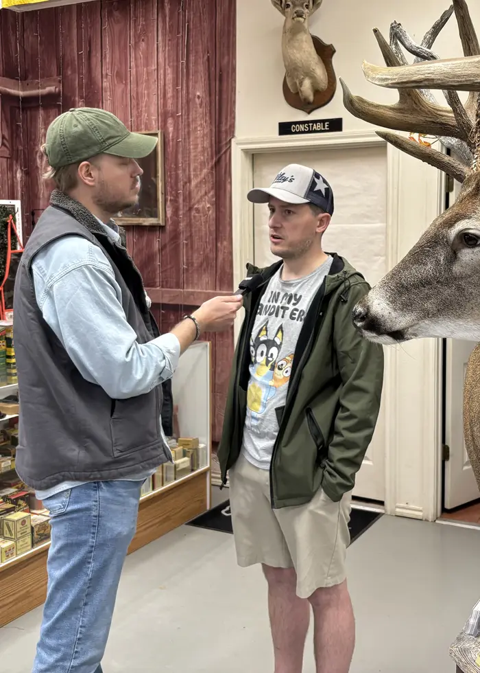 En person står i en vapenbutik med gevär och andra vapen uppställda på väggen bakom. Personen bär en grön jacka och en vit keps.