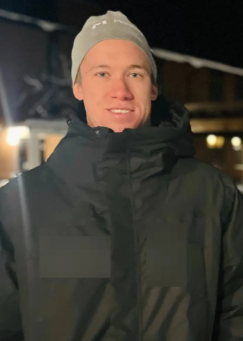 En person klädd i mörk jacka står utomhus på en snöklädd plats med belysning och byggnader i bakgrunden under kvällstid.