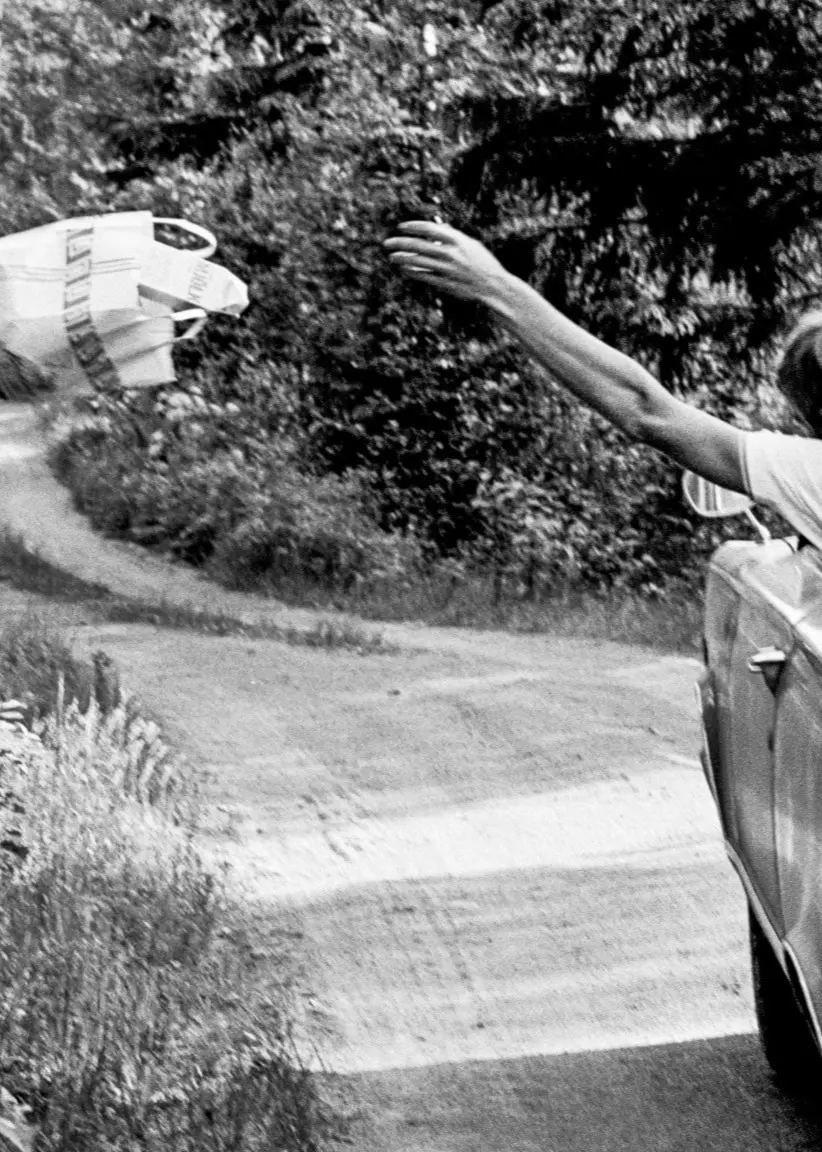 En person åker bil på en landsväg och kastar ut en påse med skräp genom bilfönstret. 