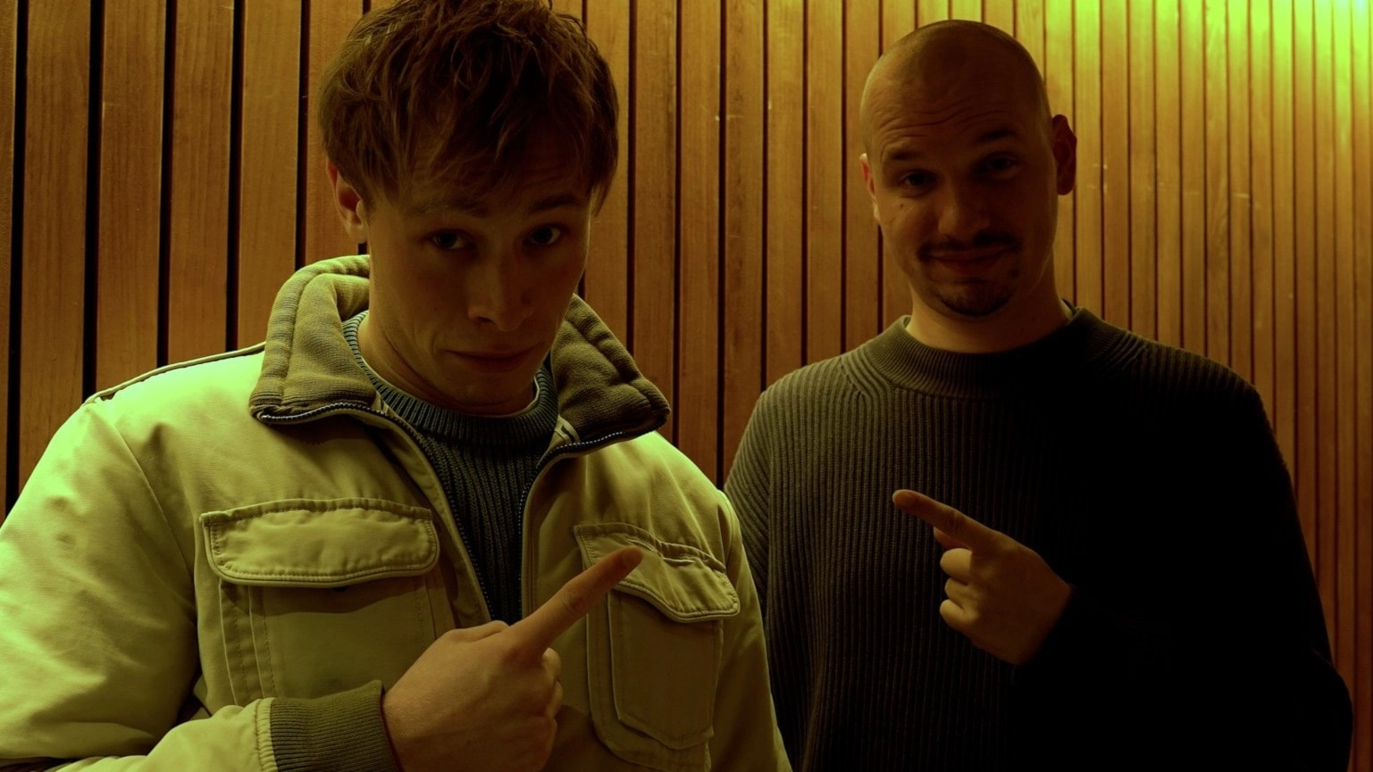 Pontus och Petter poserar framför en träpanelvägg med varmt gul belysning. Den ena bär ljus jacka, den andra mörk tröja, och båda gör pekande gest mot kameran.