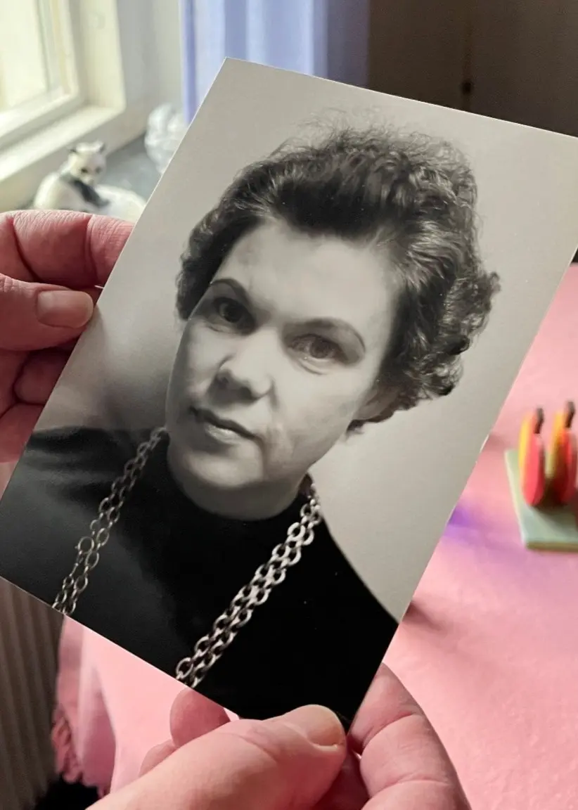 En person håller ett svartvitt fotografi med en kvinna i fokus. Bilden tas vid ett bord med rosa duk, samt fönster i bakgrunden som släpper in dagsljus.