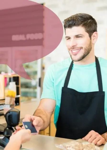 En person i blå t-shirt och svart förkläde står bakom en kassadisk i en butik och håller i ett betalningskort. En kund i randig tröja står framför disken. Miljön är ljus med trädetaljer. Artighet. Artighetsfraser.