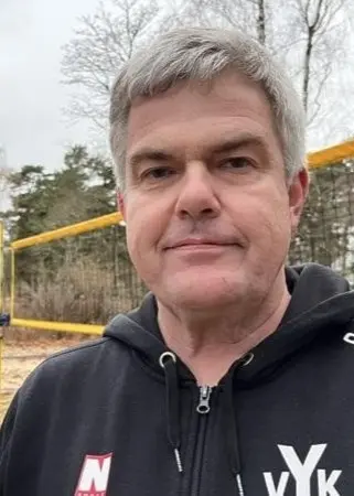 Jonas Larsson, ordförande i Ystad volleybollklubb, står framför en sandplan med volleybollnät och träd i bakgrunden, iklädd svart hoodie med klubbens logotyp.