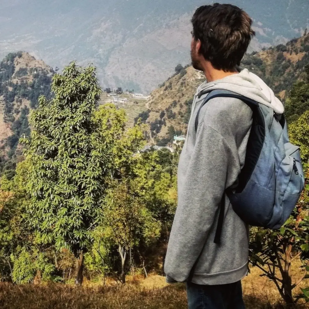 En person med ryggsäck står vänd mot en bergig och skogsklädd dalgång under en klar himmel, omgiven av grönska och majestätiska berg i fjärran.