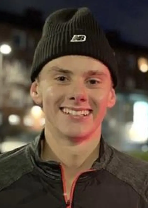 En person med mössa och sportjacka står utomhus på en nattlig stadsgata med upplysta lägenhetsbyggnader och grönska i bakgrunden.