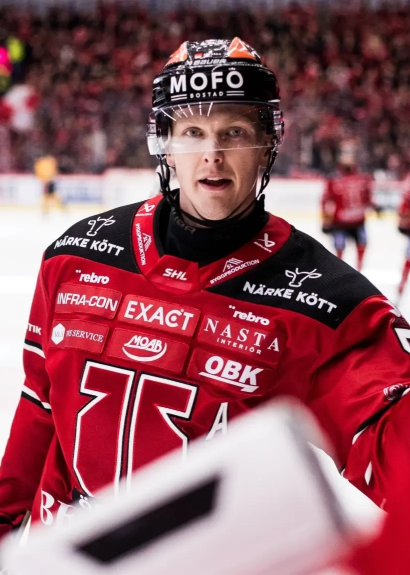 Ishockeyspelare i röd matchtröja från Örebro HK står på isen under en match, omgiven av lagkamrater och publik i bakgrunden.