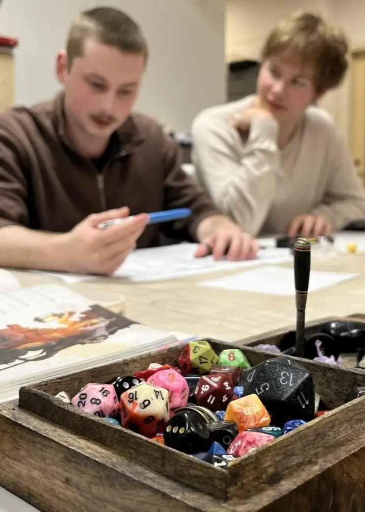 Personer deltar i ett rollspel vid ett bord med tärningar och spelmaterial. Till höger en bild på en person med en namnbricka runt halsen, iklädd en grön tröja och gul halsbandssnodd.