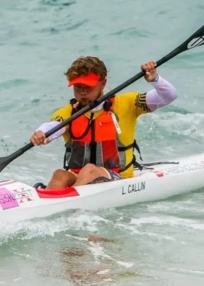 En kanotist i gul flytväst paddlar på vågigt vatten under en tävling, med en vit kanot märkt med ICF Canoe Ocean Racing-logotypen.