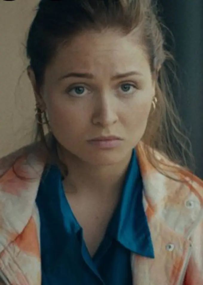 En person med uppsatt hår sitter framför en mörk bakgrund. Personen bär en blå skjorta och en ljus jacka med mönster. Bakgrunden inkluderar en suddig detalj av en möbel eller dekoration.