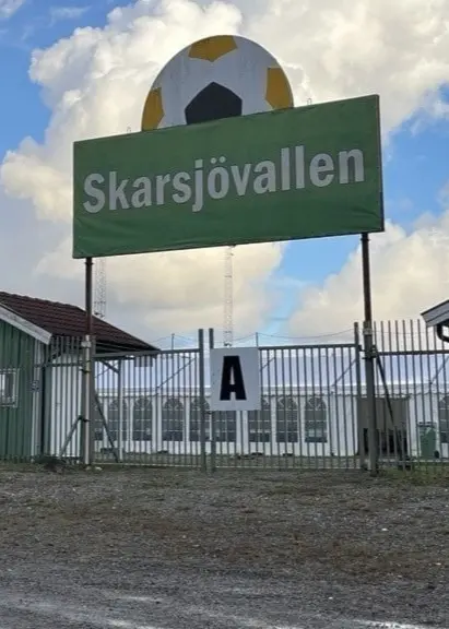 En bild på en stor grön skylt där det står ”Skarsjövallen”. Det är entréskylten till en fotbollsplan. 