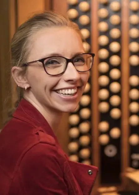 Organisten Anna Lapwood sitter vid en orgel i en röd jacka och ler. 