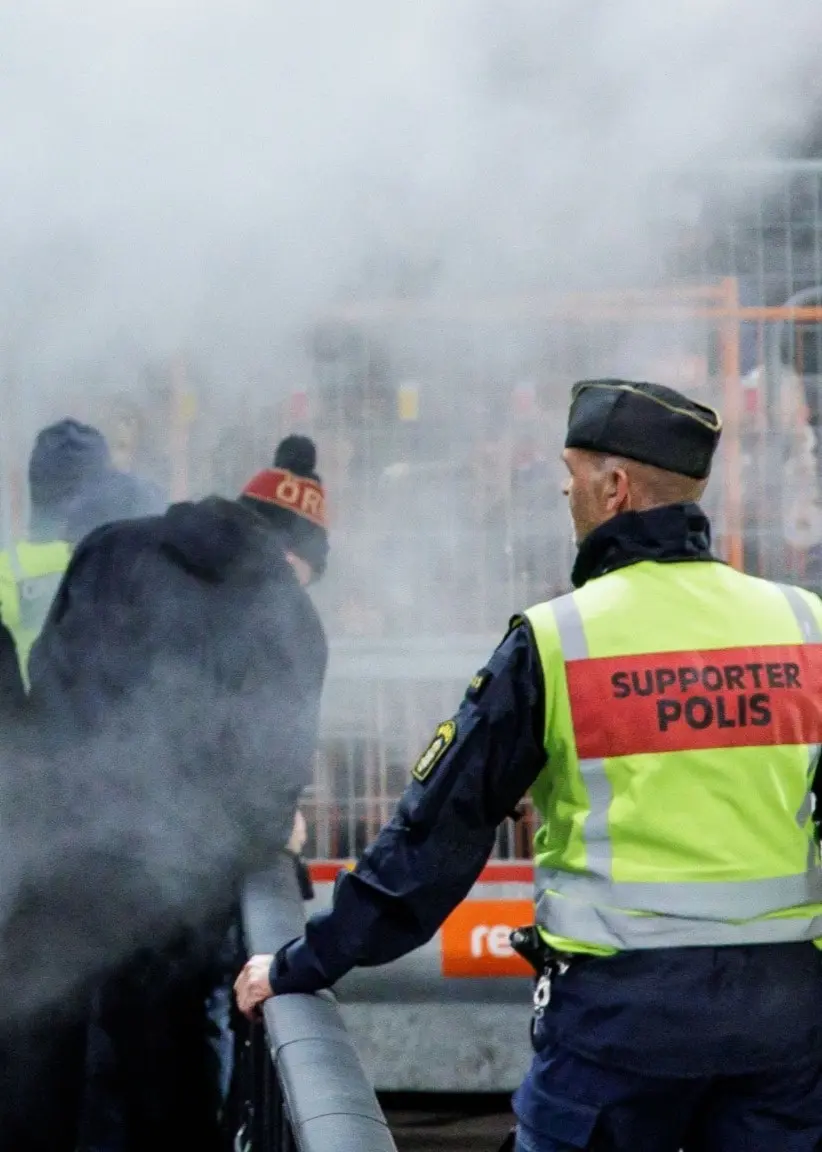 Tumult vid en fotbollsmatch mellan IFK Norrköping och Örgryte där brandsläckare används mot Örgrytes supportrar. Säkerhetspersonal i reflexvästar försöker hantera situationen.