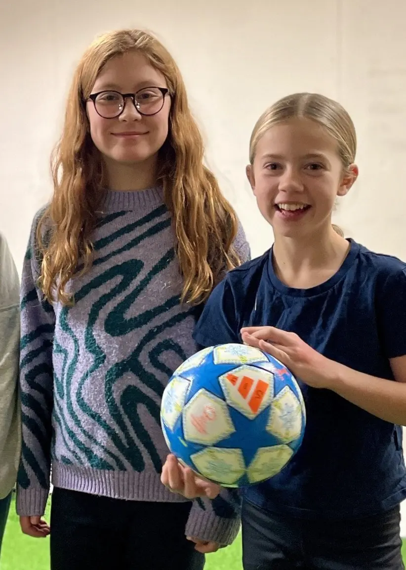 Tre unga personer, Signe, Minou och Edith, står inomhus på en konstgräsmatta. En av dem håller en fotboll.