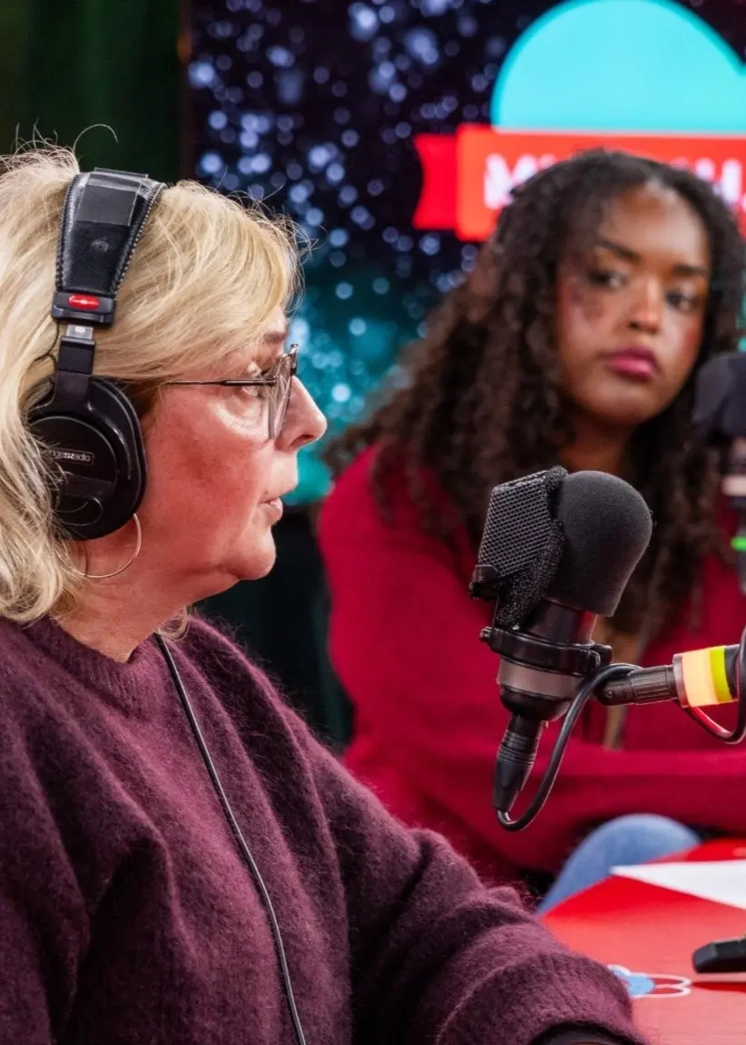 Lena Forsgren från UNICEF deltar i en radiosändning för Musikhjälpen. Hon sitter vid ett rött bord med mikrofoner, hörlurar och i bakgrunden syns en av programledarna, Assia Dahir, och Musikhjälpens logotyp.
