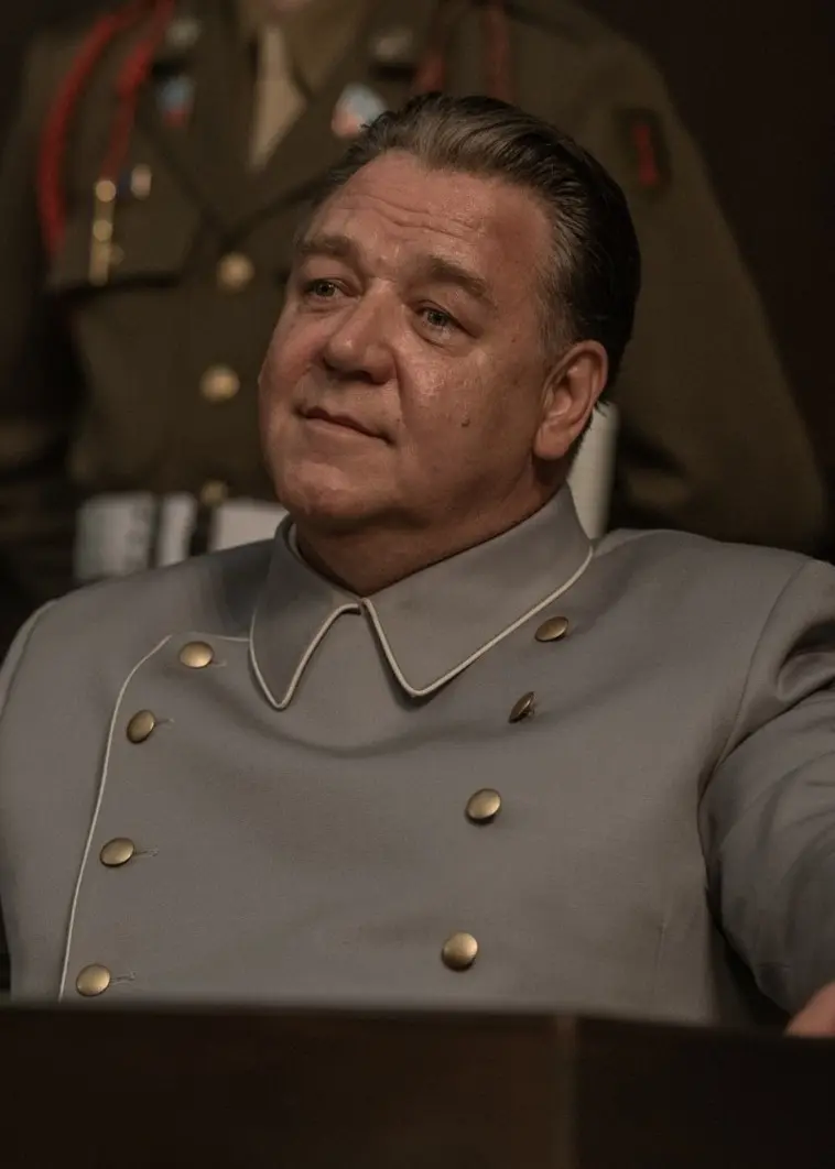 En person i grå uniform sitter vid ett bord med en mikrofon i en rättssal. En militär i uniform står i bakgrunden. Scenen förmedlar en känsla av auktoritet och formell atmosfär.