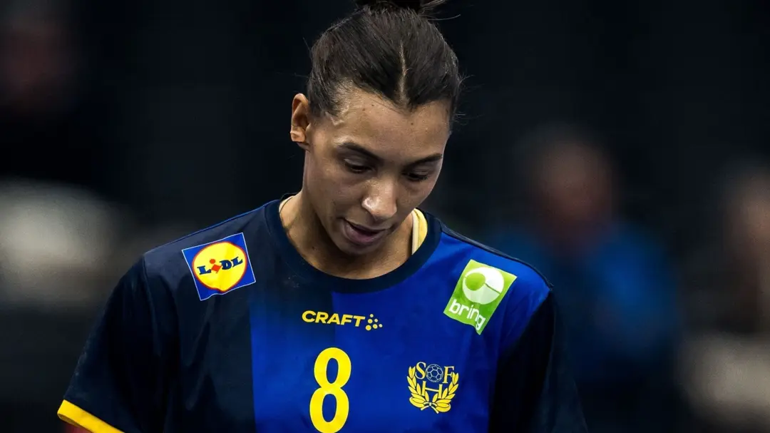 En handbollsspelare i blå och gul matchtröja med nummer 8 syns på planen. Tröjan har sponsorer som ATG och Lidl samt Sveriges emblem. Bakgrunden är mörk med suddiga konturer av publiken.