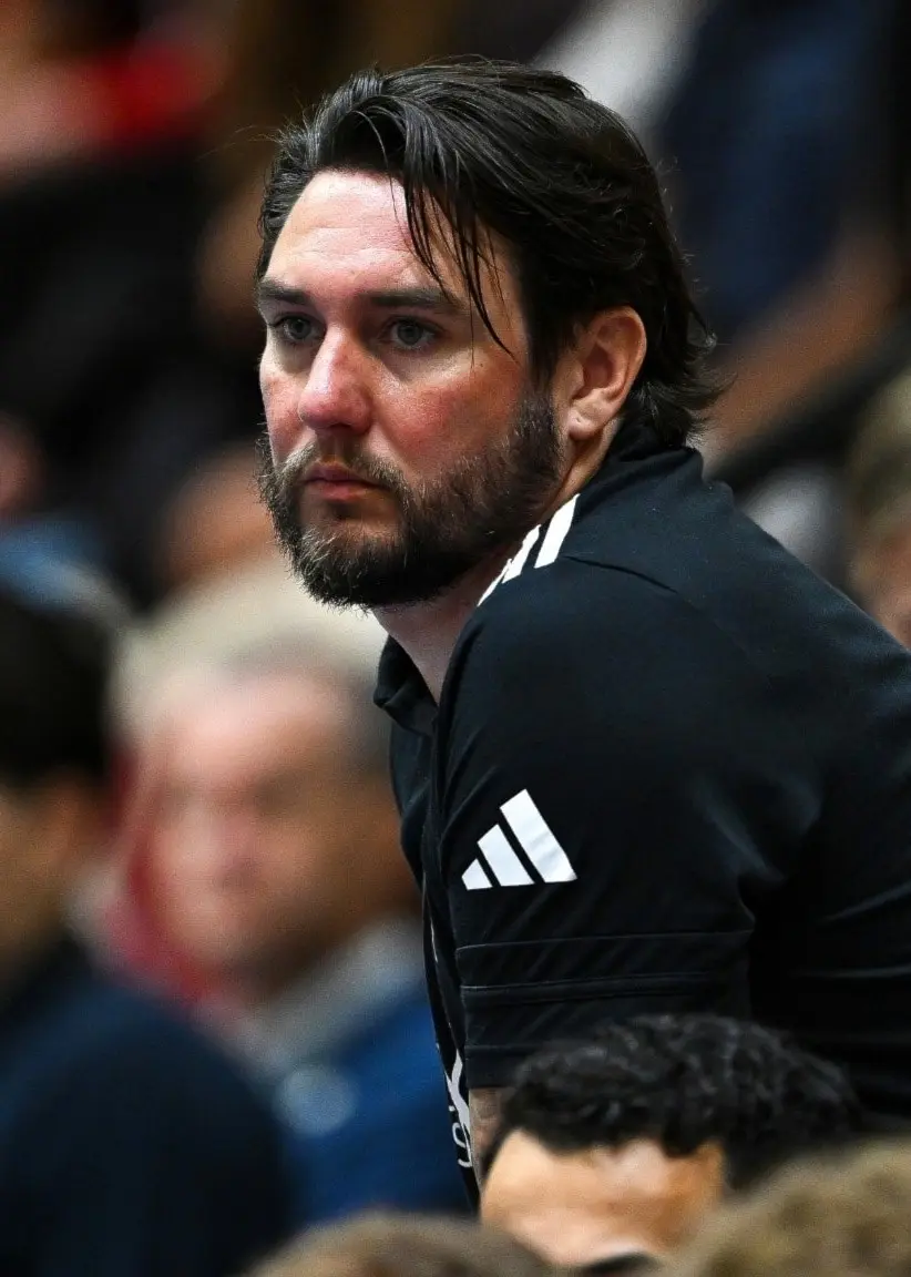 Johan Jönsson, tidigare tränare för Strängnäs IBK, ses i en innebandyhall med svart tränartröja. Han står lutad mot en barriär under en match med åskådare i bakgrunden.