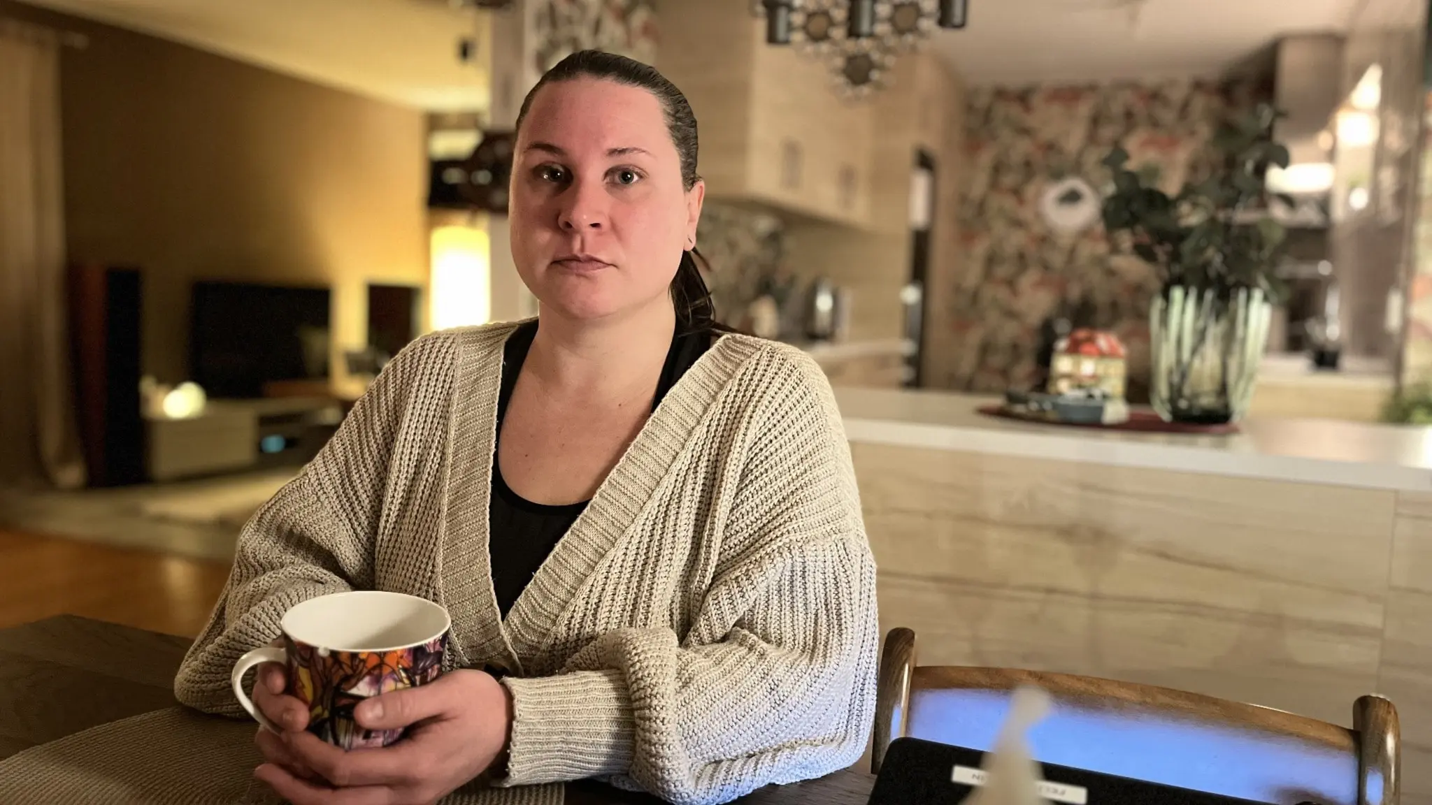 Felicia Sjölin sitter vid ett köksbord i ett modernt inrett hem, håller en kopp och bär en ljus stickad tröja. I bakgrunden syns en öppen köksmiljö med dekorativa inslag.