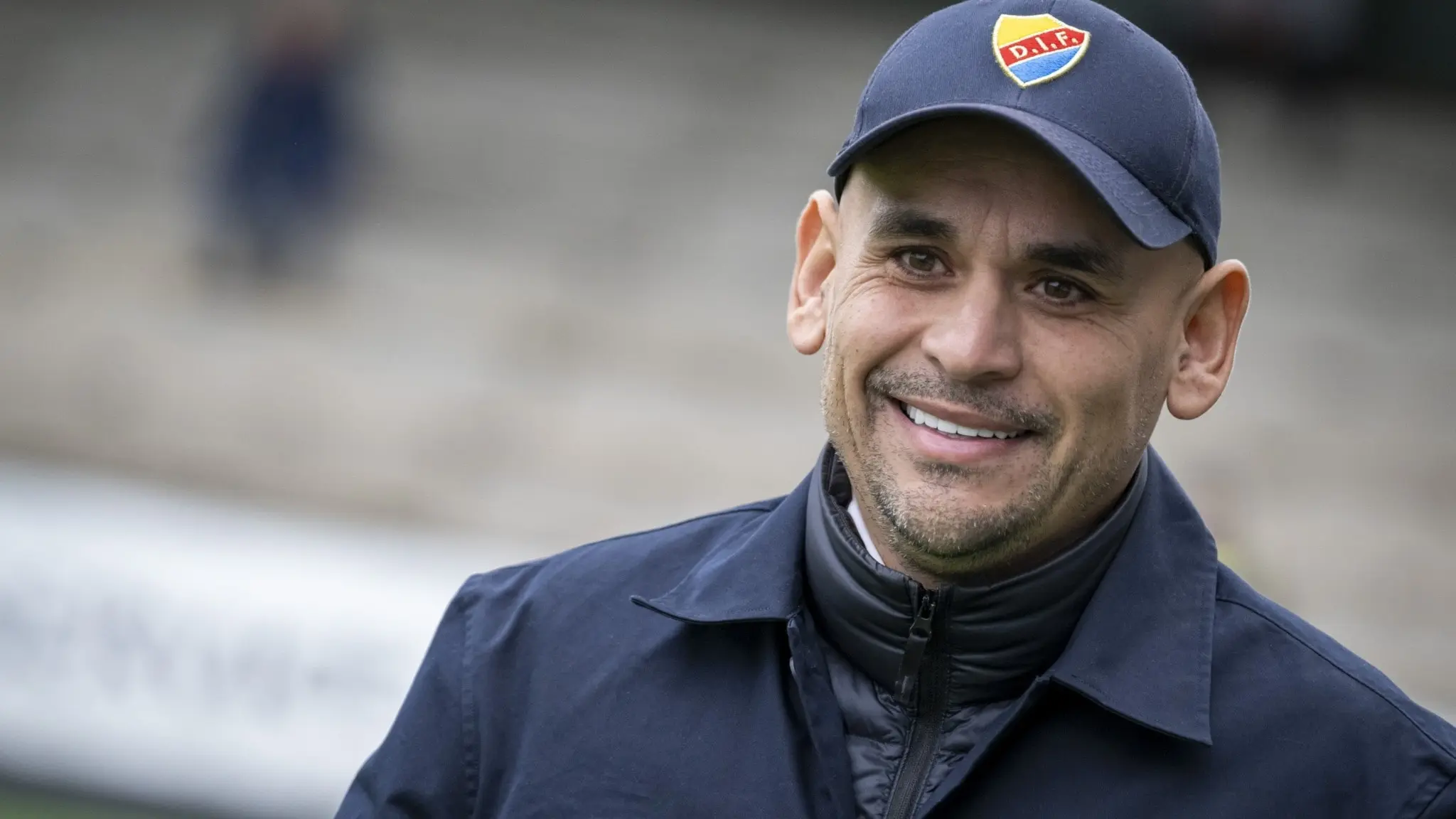 Djurgårdens tränare Marcelo Fernández står med klubbens logotyp på keps och jacka inför matchen mot FC Rosengård i damallsvenskan på Malmö IP. Bakgrunden visar en del av läktaren.