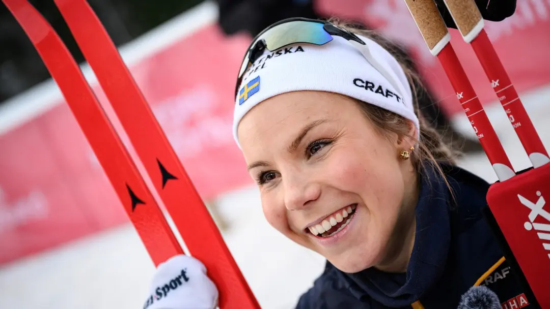 Johanna Hagström ler och håller i sina skidor efter en världscupseger i Trondheim. Hon bär skidglasögon och pannband med snö och en röd skylt i bakgrunden.