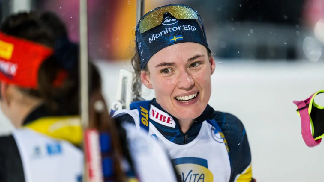 En tävlingsåkare i skidkläder, sponsrad av Vita, syns med skidor och stavar i ett sportevenemang. Personen står på en snötäckt bana omgiven av andra deltagare med tävlingsutrustning.