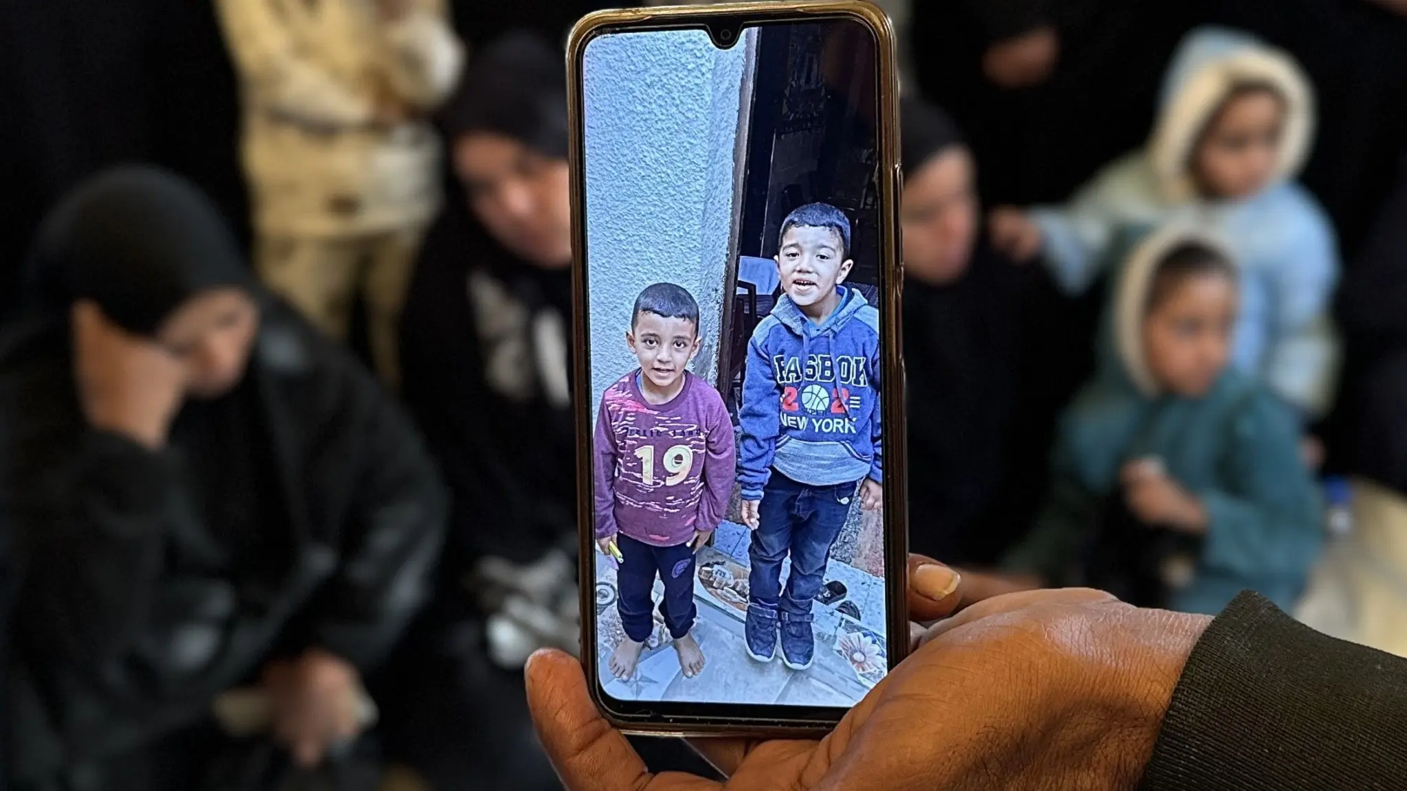 En person håller upp en mobiltelefon som visar två barn som dödades av israeliska drönare i Gaza. Sörjande personer sitter i bakgrunden.