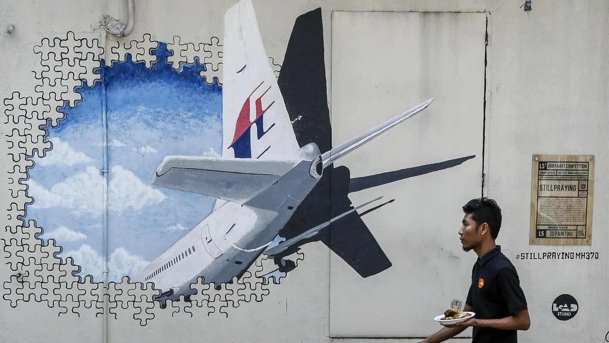 En väggmålning i Shah Alam, Malaysia, visar ett Malaysia Airlines MH370-flygplan som verkar krascha genom en vägg mot en blå himmel. En man med en bricka passerar målningen.
