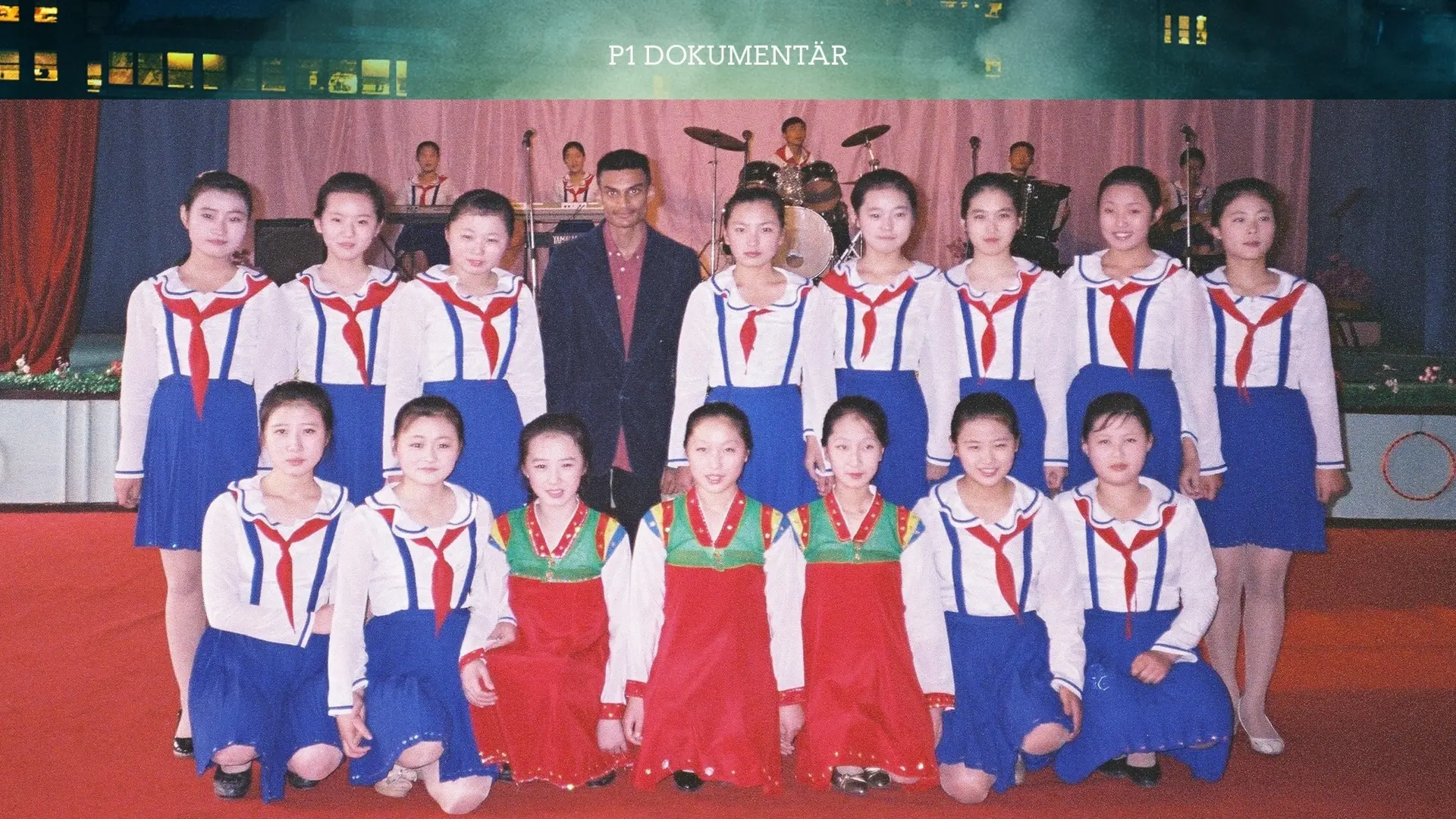 15 flickor i blo-vita dräkter står uppställda som på en klassbild. I mitten av den översta raden står Robin Jonsson med vinröd skjorta och mörk kavaj.