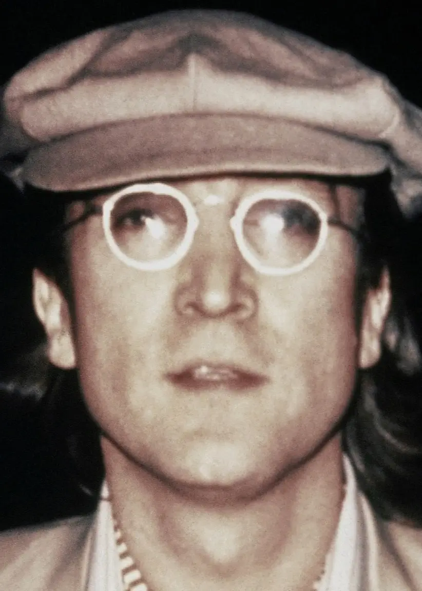 John Lennon ser allvarlig ut. En närbild på hans ansikte. Han har runda glasögon och en bär en keps.