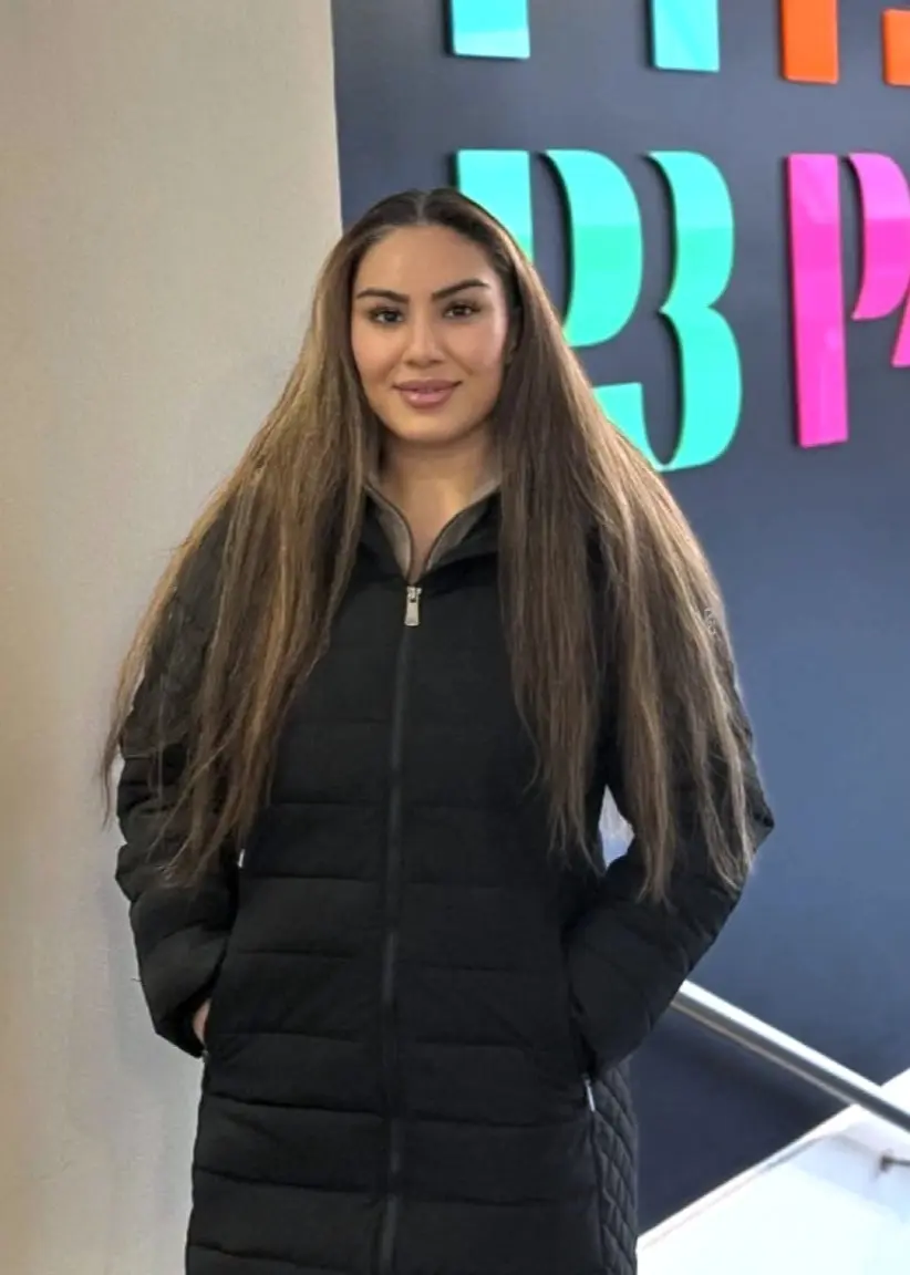 En bild som visar Migrationsverkets logotyp på en byggnad till vänster och en kvinna i svart jacka med långt hår framför en färgglad vägg till höger.