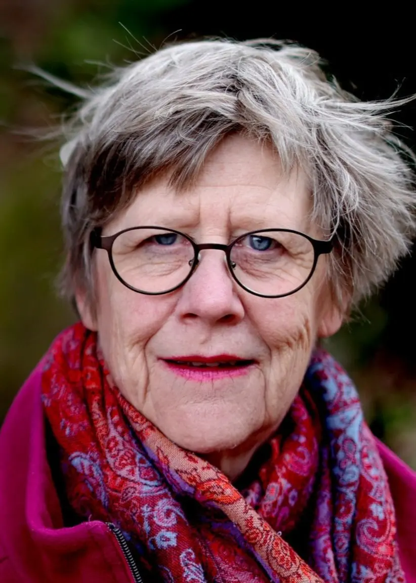Agnes Wold, svensk läkare och professor i klinisk bakteriologi, står lutad mot ett träd i en skogsmiljö. Hon bär en rosa kappa och en färgglad halsduk.