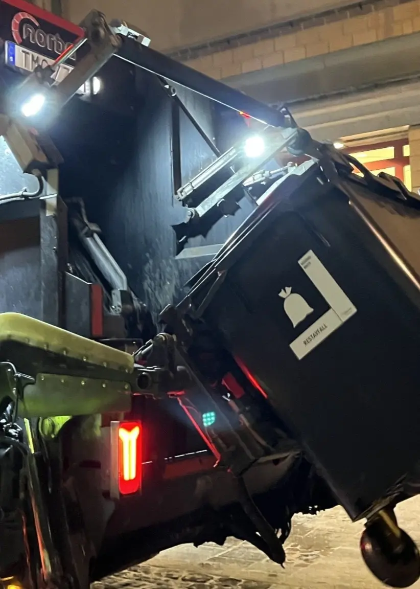 Två soparbetare i gula reflexkläder arbetar vid en upplyst sopbil och tömmer soptunnor på en stadsgata under tidig morgon. Boende i centrala stan har klagat att de blir väckta i ottan av ljudet från sophämtningen i en namninsamling.