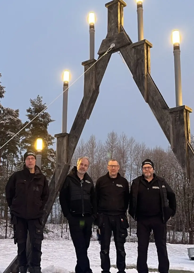 Fyra personer står framför Lagans stora ljusstake som lyser upp i vinterlandskapet. Ljusstaken är en imponerande konstruktion med flera tända ljus, omgiven av snö och skog.