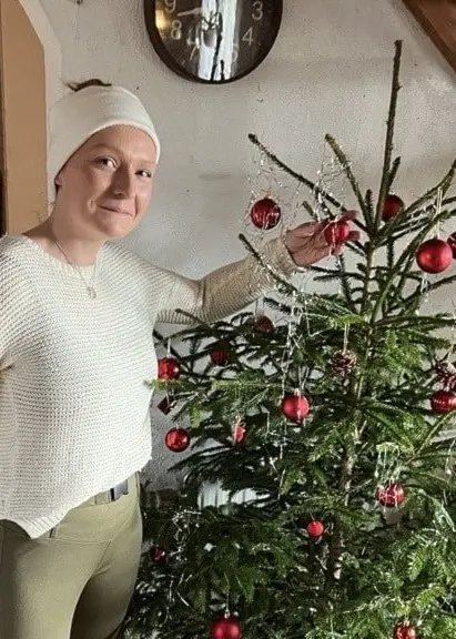 Pamela Freborn står bredvid en julgran dekorerad med röda kulor i ett träinrett hem. Hon berättar att hon ger granen pilsner för att hålla den livfull och fin från november till tjugonde knut.