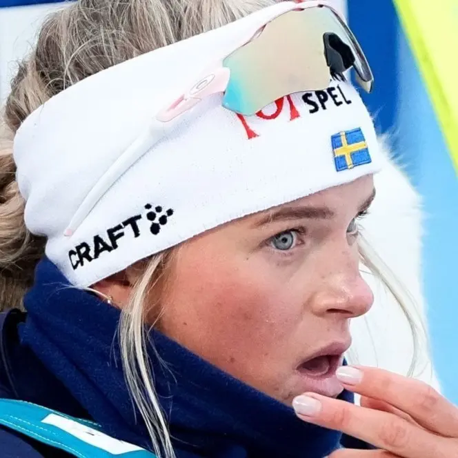 Fyra längdskidåkare tävlar i en snöklädd skog under en internationell skidtävling. De är klädda i färgglada tävlingsdräkter med startnummer, och skidspåret är omgiven av vinterlandskap.