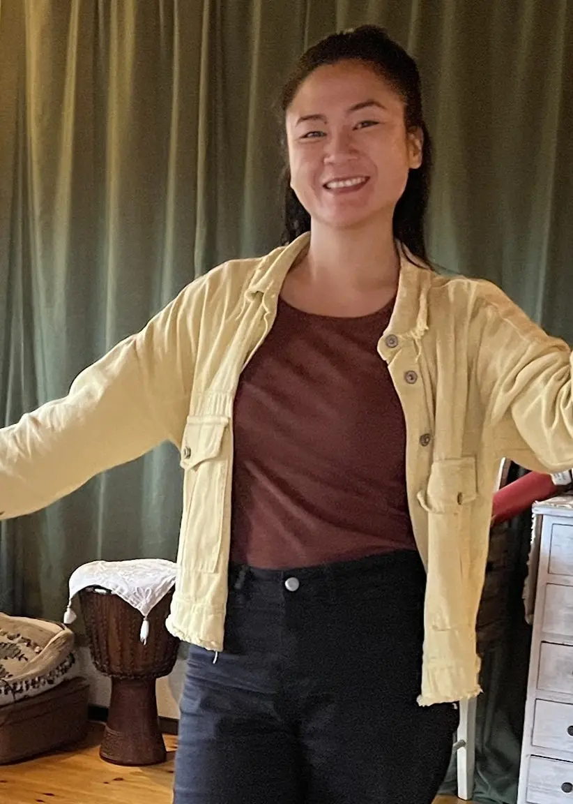 En kvinna, Mai Huynh, står i ett vardagsrum med trägolv och gröna växter, klädd i gul skjorta och mörka byxor, omgiven av en inredning med gardiner, mattor och möbler.