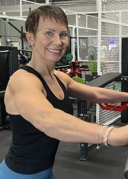 Nina Olli tränar på gymmet med hjälp av träningsmaskiner, klädd i träningslinne och tights. Miljön är modern med flera gymmaskiner och nätväggar i bakgrunden.