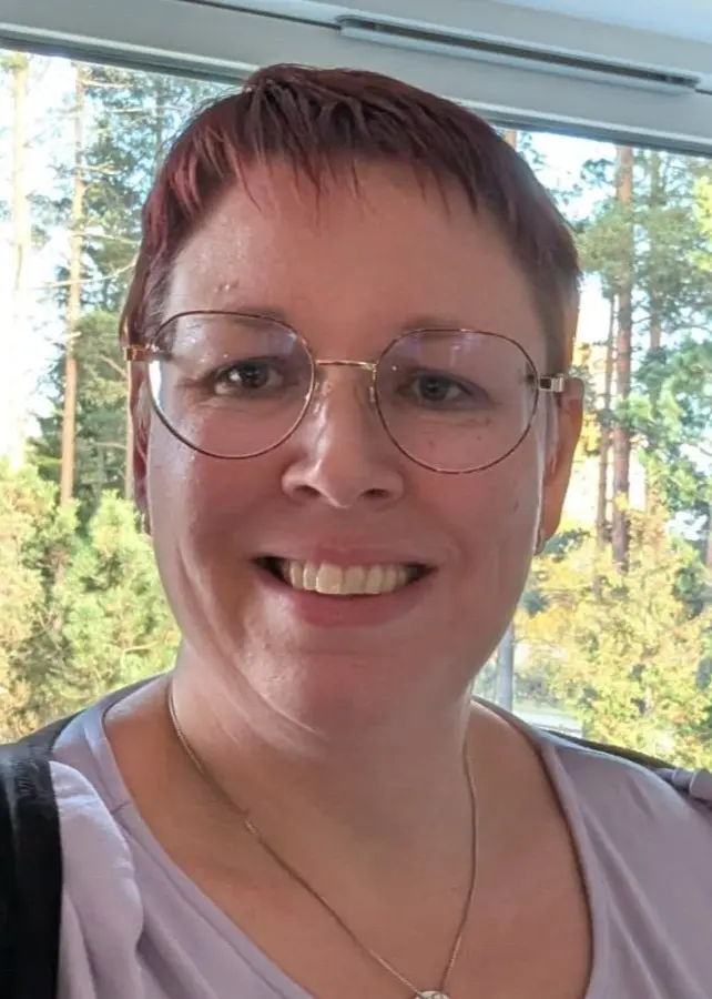 Linda Poralis, ordförande för Fragile X i Sverige, står vid ett fönster med blåmönstrade gardiner. Utanför syns grönska. Hon har en ljus topp och ett halsband.