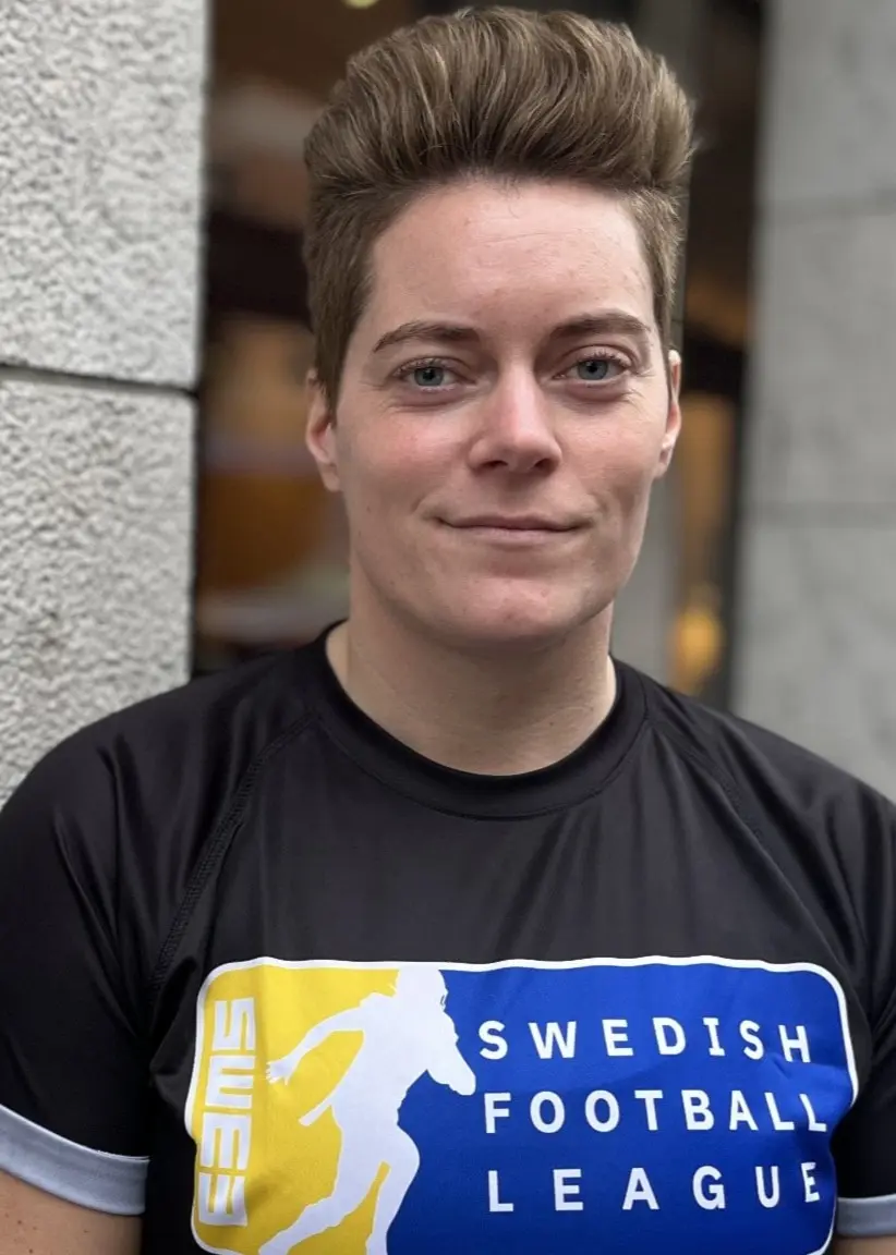 En person står vid en grå vägg och bär en svart t-shirt med texten 'Swedish Football League' och en blå-gul grafisk design på bröstet
