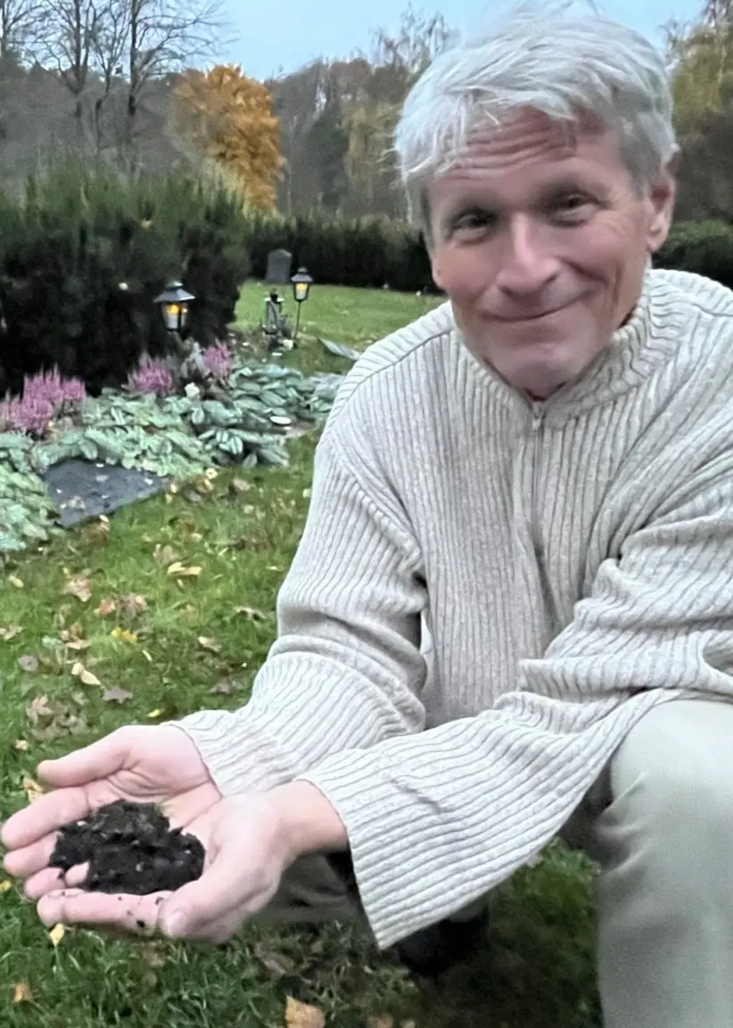 Anders Krasse från Svenljunga sitter i en park med höstlöv och gröna buskar i bakgrunden, och håller jord i sina händer för att symbolisera sin önskan att bli återjordad efter döden.