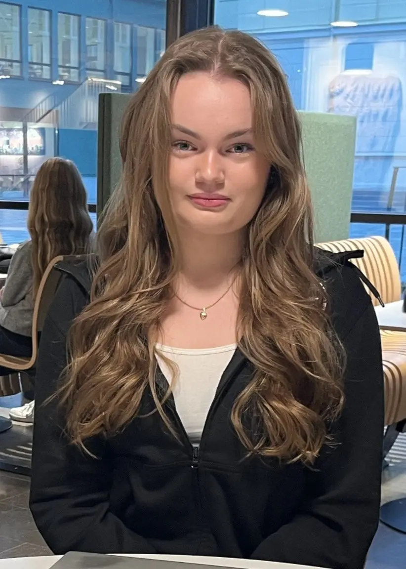 Porträttbild flicka i gymnasieålder med långt, brunt hår i ett klassrum.