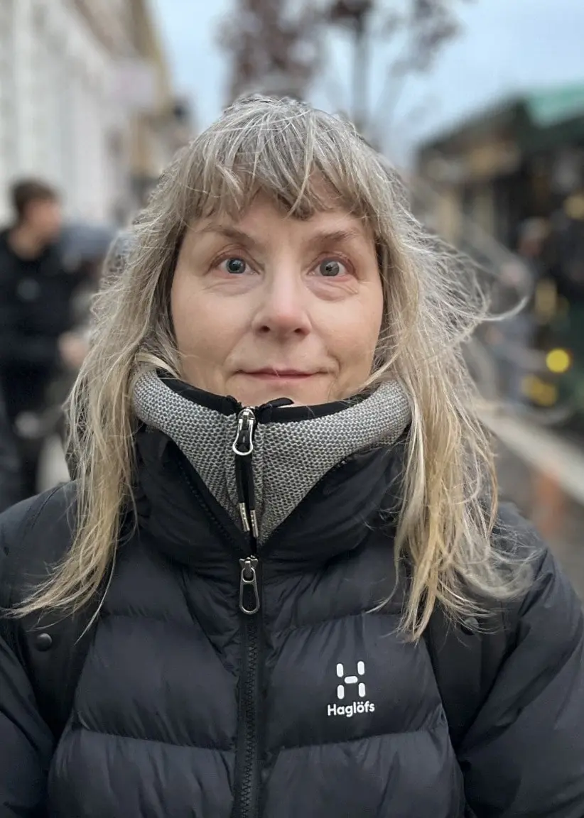 Carola Weckström står på en trottoar klädd i svart vinterjacka och halsduk, med en grön buss parkerad i bakgrunden på en stadsgata.